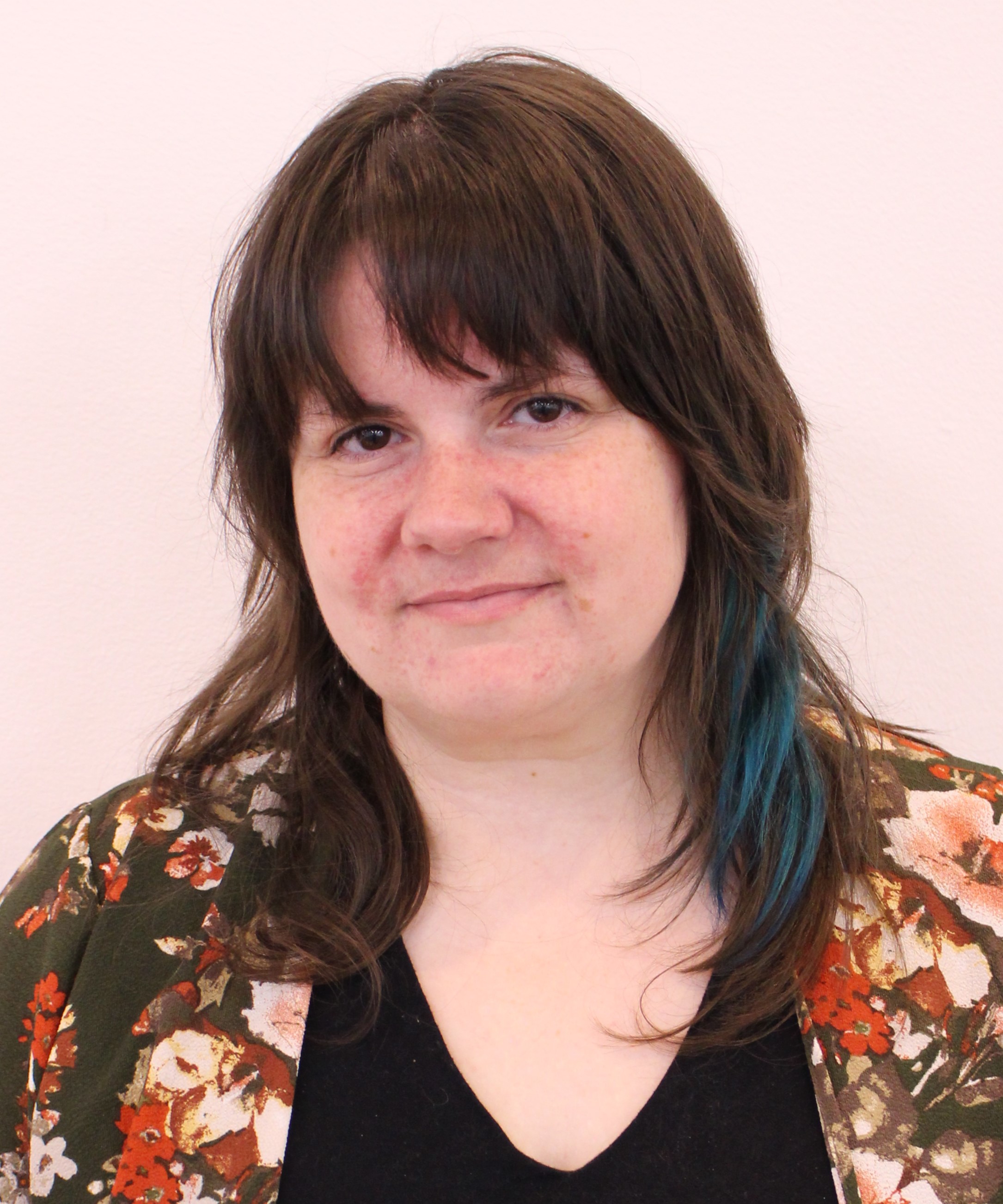 A headshot of CCJPL's tween librarian, Megan Heyl. She has blue highlights in her hair and sits in front of a plain white background while wearing a colorful shirt